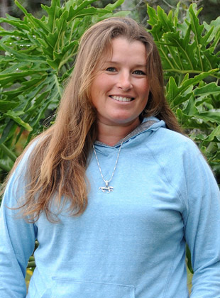 A person in a blue hoodie smiling, standing in front of green foliage.